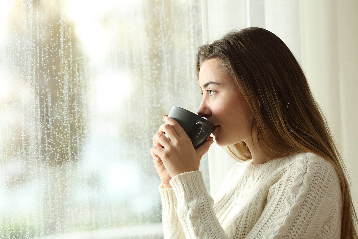 Teen drinking coffee looking through a window a rainy day
